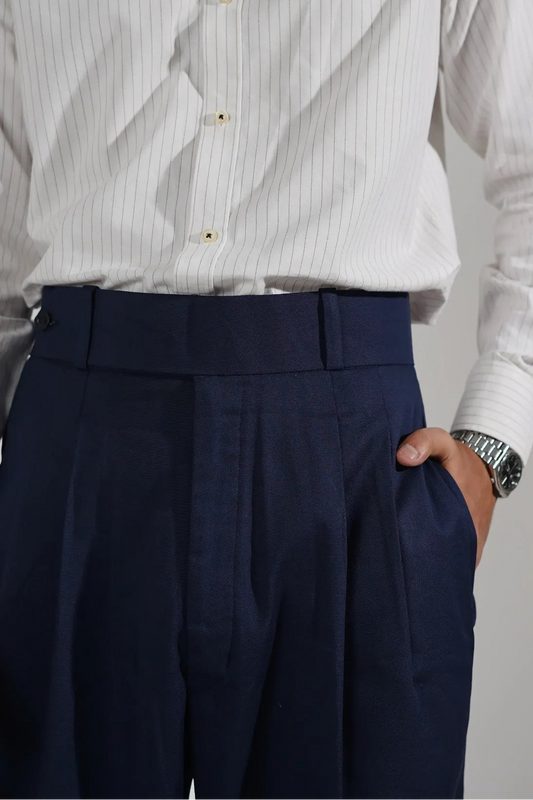 A close-up of a person wearing a white long-sleeve button-up shirt with thin vertical pinstripes and a small embroidered red monogram on the chest, paired with dark navy trousers. The person's hand is in the pocket of the trousers, and a silver watch is visible on the wrist, set against a plain light gray studio background.