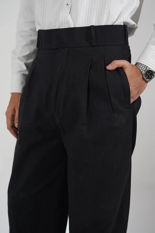 A close-up of a person wearing black pleated trousers and a white striped dress shirt, with hands holding the waistband to show the fit and fabric.