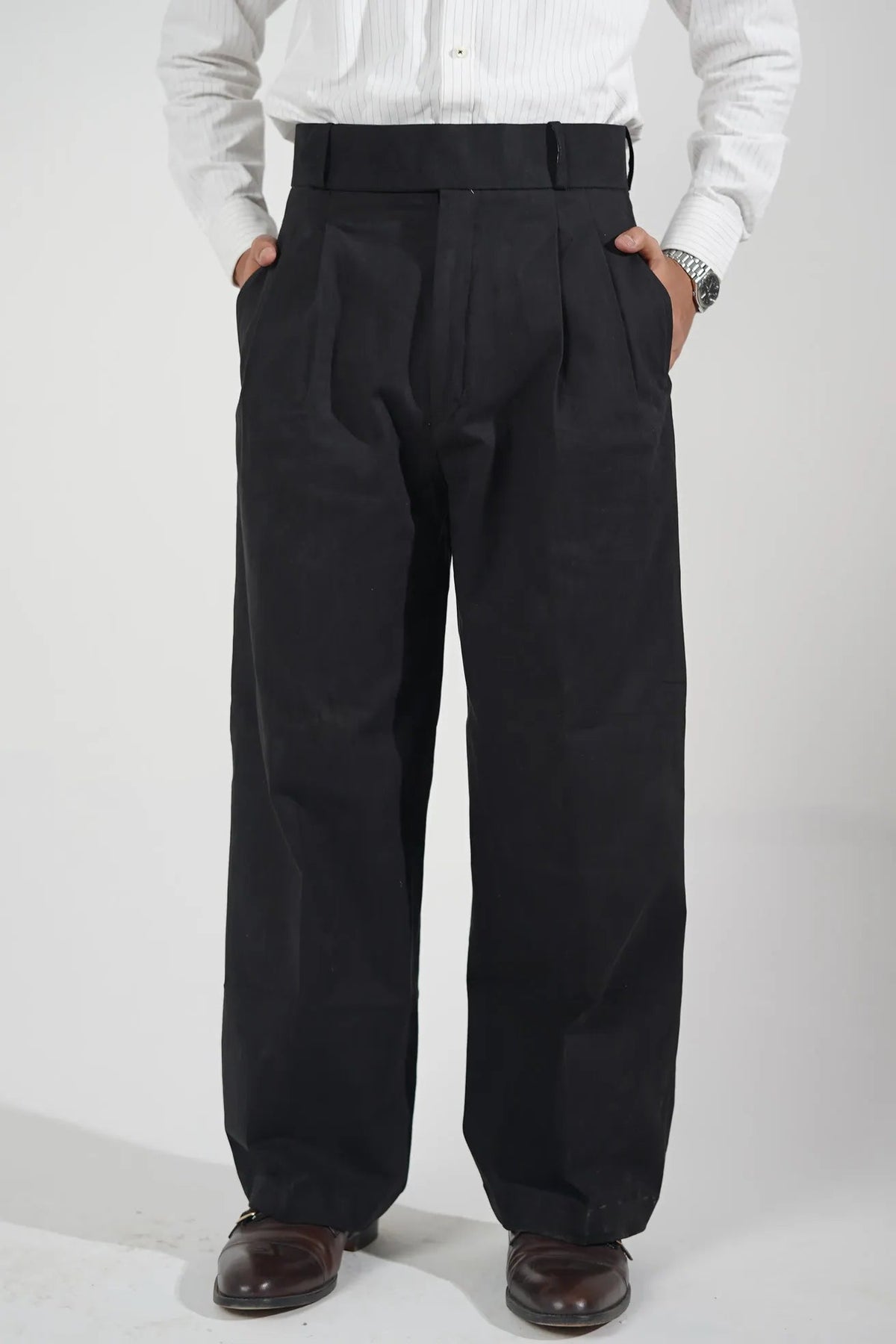 A person wearing black pleated trousers with a black belt, paired with a white long-sleeve shirt and brown leather shoes, shown from the waist down against a white background.