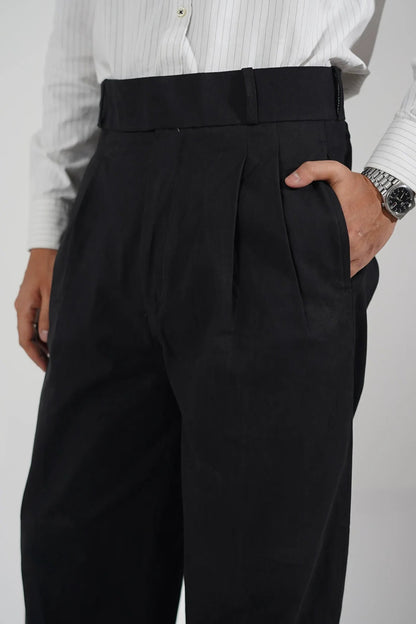 A close-up of a person wearing black pleated trousers and a white striped dress shirt, with hands holding the waistband to show the fit and fabric.