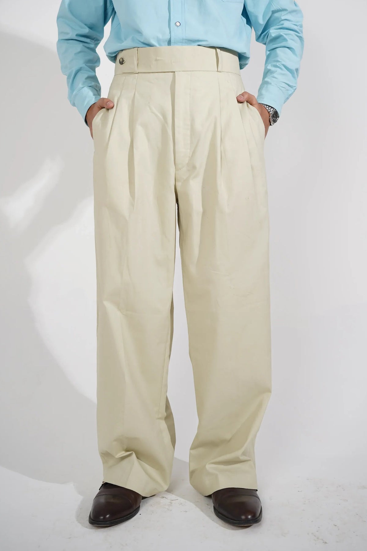 A person wearing beige wide-leg pleated trousers, a light blue long-sleeve button-up shirt, and dark brown leather shoes, photographed from a high angle against a white studio background.