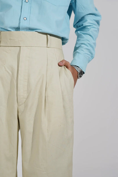 A close-up of a person wearing light beige trousers and a light blue button-up shirt, with the hand adjusting or holding the waistband of the pants against a plain light gray background.
