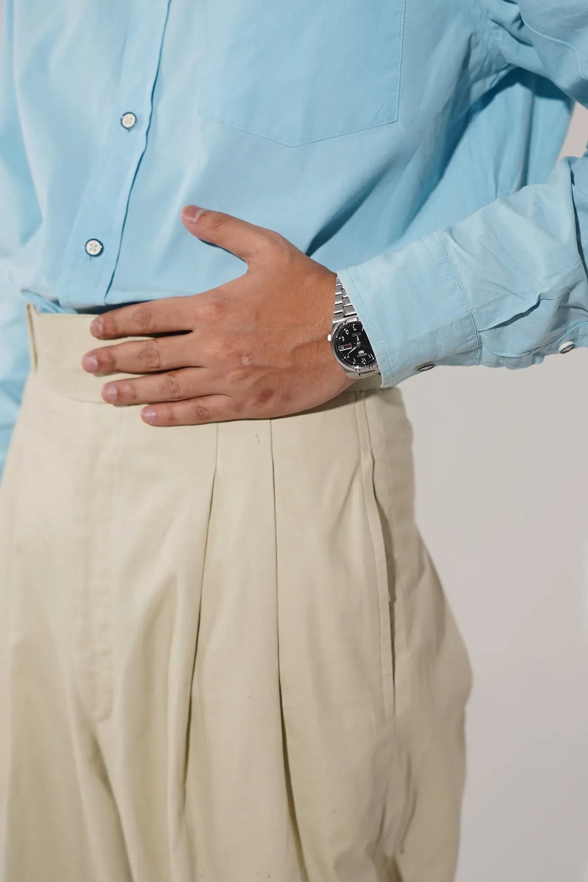 A close-up shot of a person wearing a light blue button-down shirt and beige trousers, with their hand resting on the waistline of the pants. A silver watch is visible on the wrist.