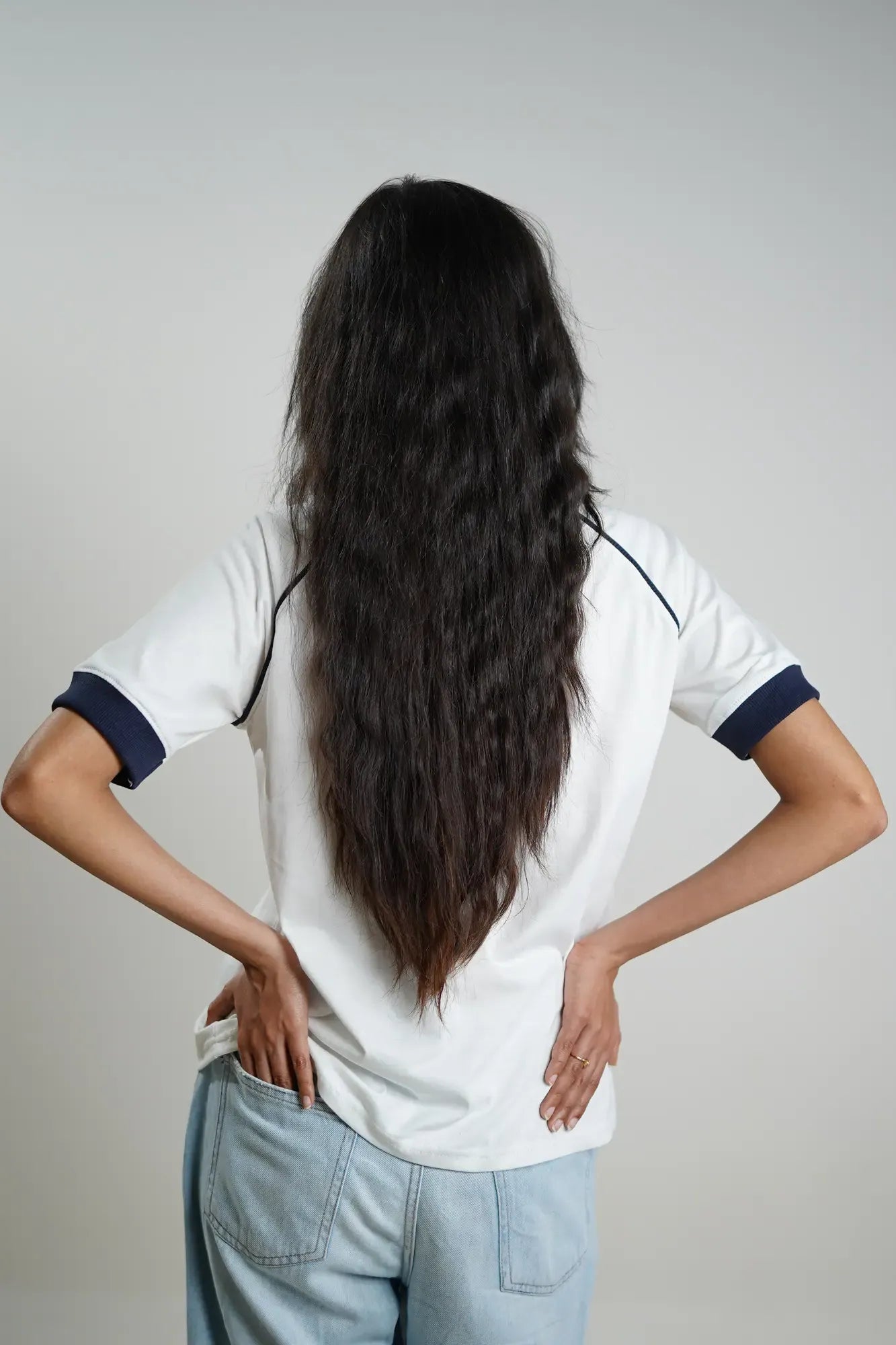 Woman wearing a white shirt with navy contrast sleeves and light blue jeans, standing with hands on hips against a plain background
