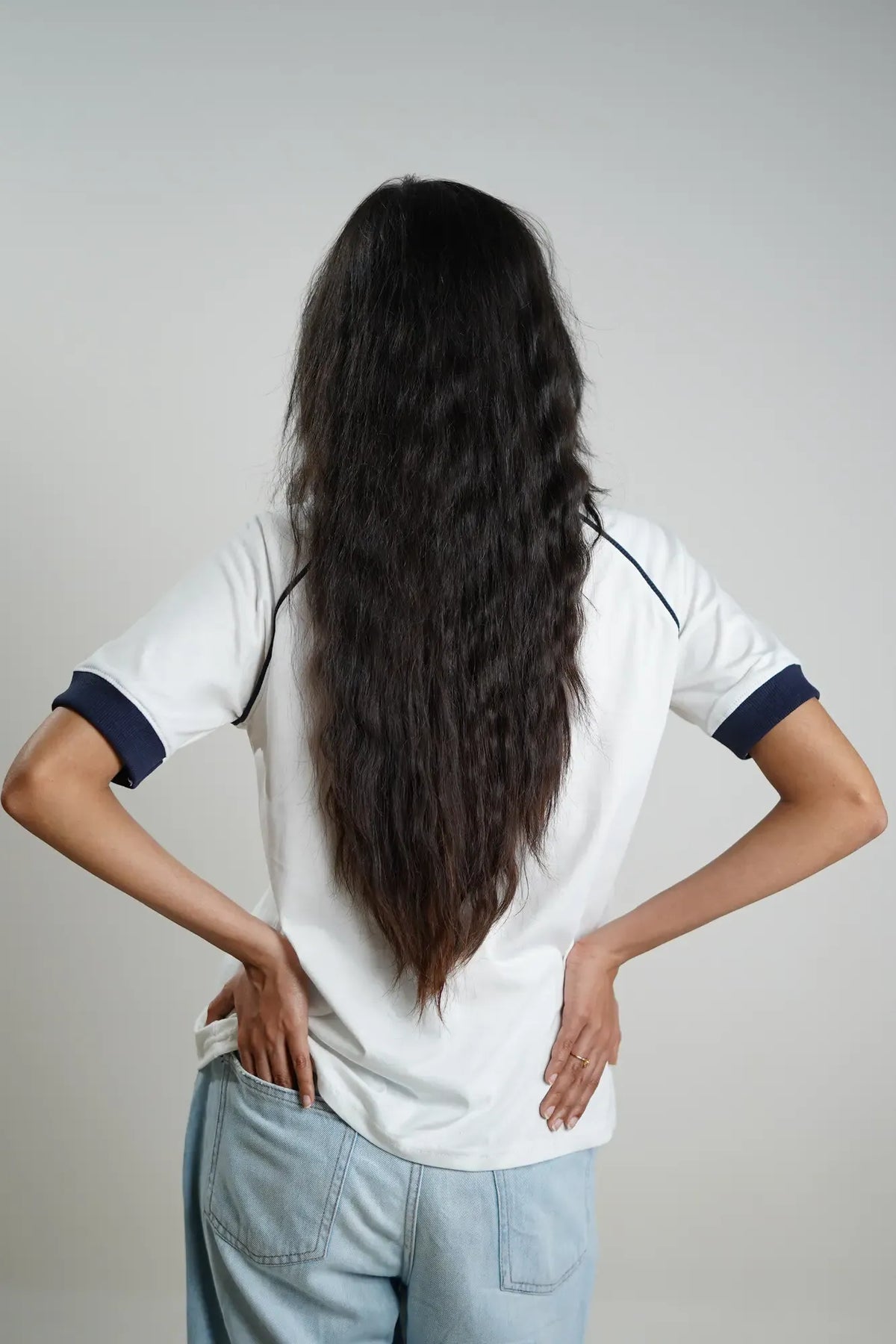 Woman wearing a white shirt with navy contrast sleeves and light blue jeans, standing with hands on hips against a plain background