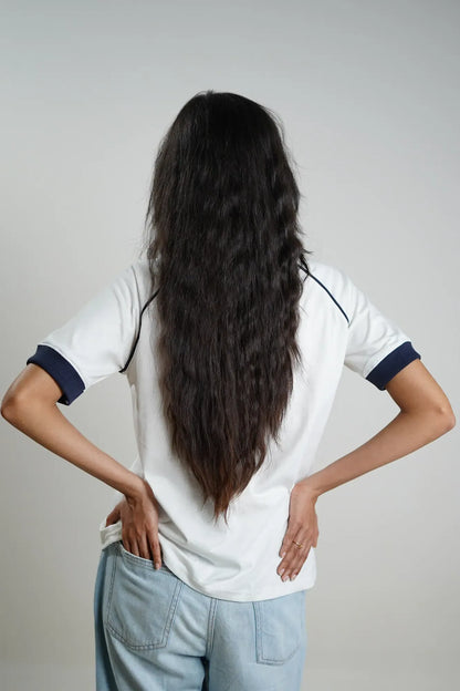 Woman wearing a white shirt with navy contrast sleeves and light blue jeans, standing with hands on hips against a plain background
