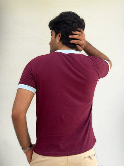 Man wearing a burgundy / maroon t-shirt with sky blue trim against a plain background