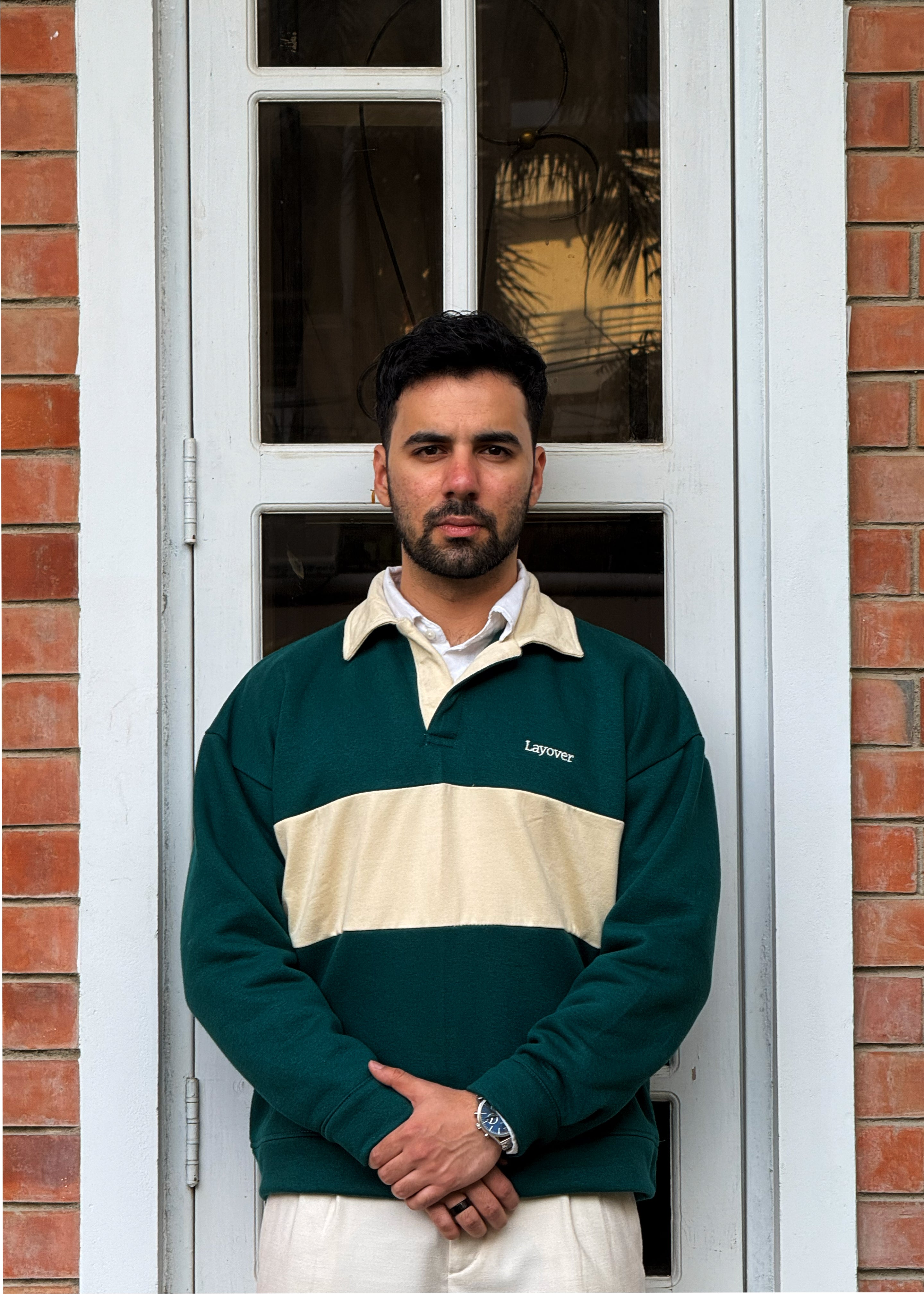 Man wearing the Layover Project's classic relaxed green and beige striped rugby sweater standing in front of a brick wall with a window. 